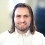 a young white man with shoulder-length brown hair, wearing a white button up shirt