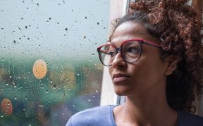 a photo of a Black woman with glasses sitting next to a window with rain splattered on the glass