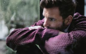 a photo of a man in a sweater looking concerned, and looking outside at the rain