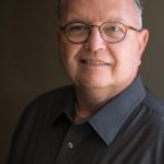 an older white man with glasses wearing a light black button-up shirt