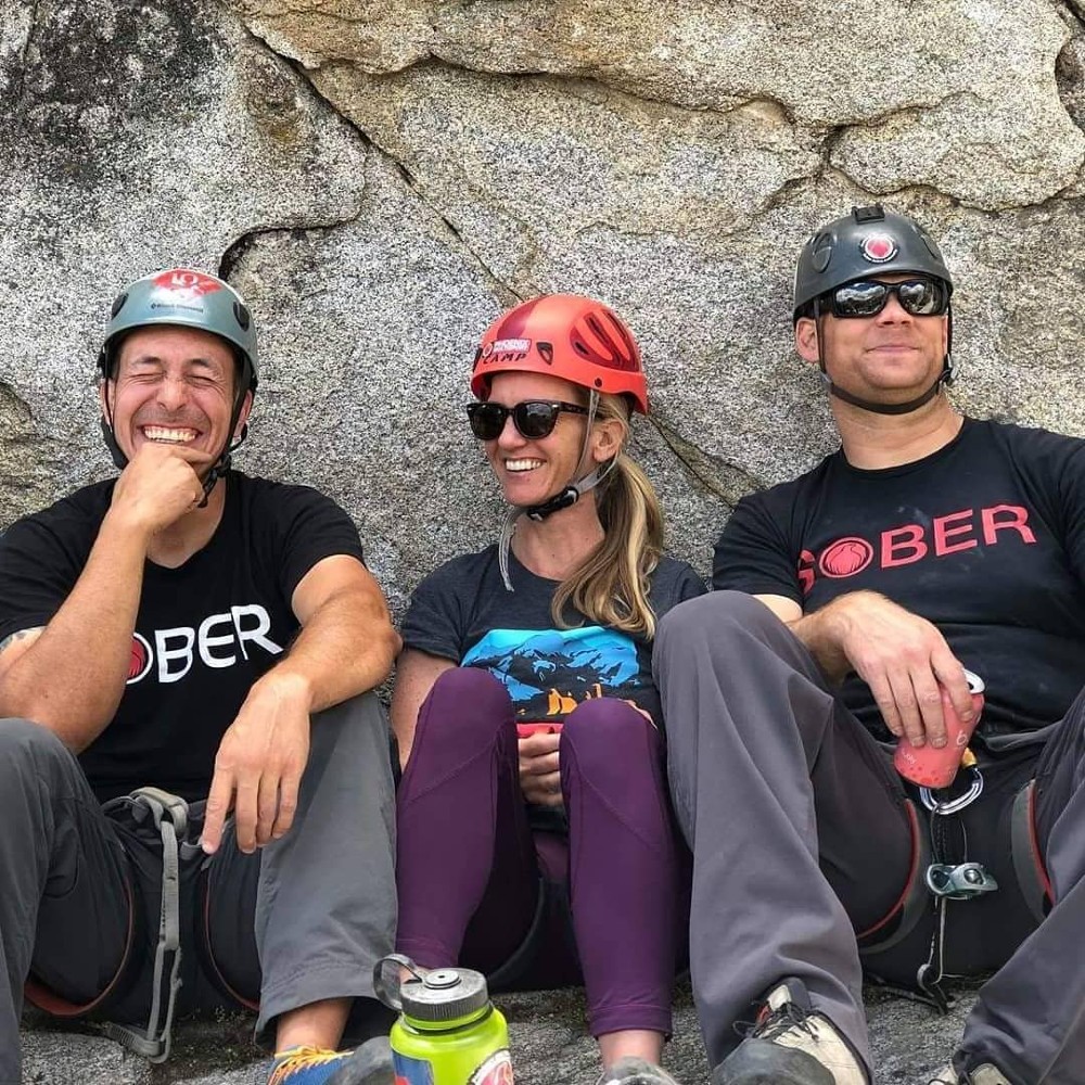 a photo of two men, one woman (all white) in climbing gear sitting in front of a rock wall