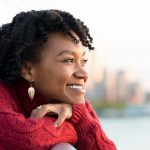 a Black woman in a red sweater in front of a water-way skyline, smiling
