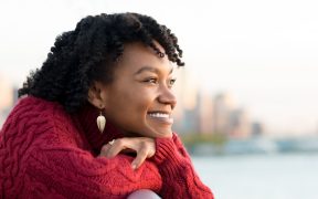 a Black woman in a red sweater in front of a water-way skyline, smiling