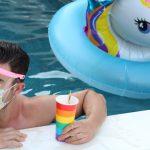 a photo of a white man at the pool, with a blow up toy next to him, wearing a face shield and mask and a drink in his hand