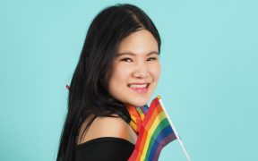 an Asian woman, black hair and wearing a black top, looking at the camera from the side, with a Pride flag in one hand