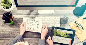 a photo of two people at a desk, one with a piece of paper on it with a website design layout on it, the other with a tablet in their hand, in front of a computer