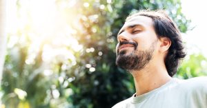 a photo of a man with a beard, dark, with his eyes closed and smiling in the sun with trees behind him