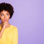 a photo of a Black woman in a yellow long sleeve shirt, with one hand on her chin, looking up in contemplation, against a purple background