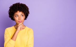 a photo of a Black woman in a yellow long sleeve shirt, with one hand on her chin, looking up in contemplation, against a purple background