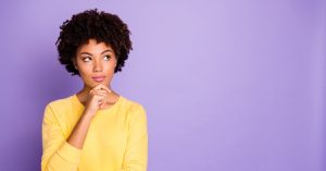 a photo of a Black woman in a yellow long sleeve shirt, with one hand on her chin, looking up in contemplation, against a purple background