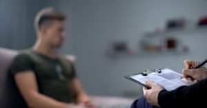 a photo of a person with a notebook and pen looking at a blurred image of a man in a t-shirt on a couch