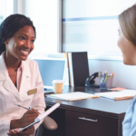 a black woman in a doctor's coat sits on the left, ready to write something. A white woman in brown hair sits across from her