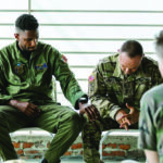 A group of men sitting in a circle, all wearing military fatigues. One man has his hand on the knee of the other in comfort