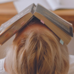 a person with blond hair and white tshirt laying down with a book on their face