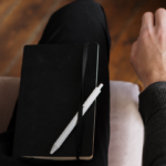 a closeup of a notebook and pen on the right leg of a lap of someone sitting in a chair