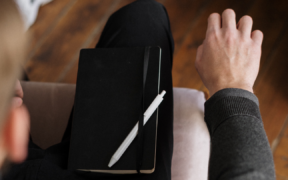 a closeup of a notebook and pen on the right leg of a lap of someone sitting in a chair