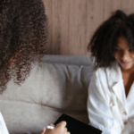 the back of an African American with a white shirt, talking to another African American on a couch