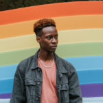 a black person with short hair wearing a jean jacket and light shirt standing in front of a wall with the pride flag on it