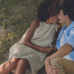 a Black couple, one in a sundress and the other in brown slacks and a blue top, sit on the grass kissing
