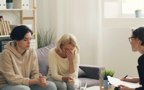 two women on a couch, one with black hair in a hoodie and jeans and the other with blonde hair and a yellow top and blue jeans. The blonde has her hand over her eyes. Across from them sits a woman with black hair wearing a black suit, holding a pen and pad of paper