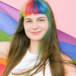 a white young person with hair with streaks of color, wearing a white tank top, holding a pride flag.