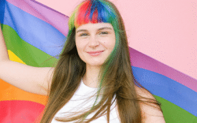 a white young person with hair with streaks of color, wearing a white tank top, holding a pride flag.