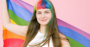 a white young person with hair with streaks of color, wearing a white tank top, holding a pride flag.