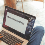 a white person of indistinguishable gender has a laptop open on their lap (wearing jeans0. The laptop says 'marketing strategy'