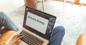 a white person of indistinguishable gender has a laptop open on their lap (wearing jeans0. The laptop says 'marketing strategy'