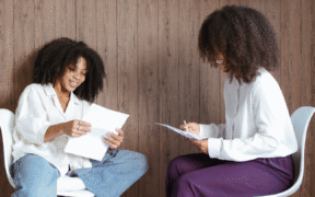 Two black women sit in white chairs, holding papers. The one on the left wears a white shirt and blue jeans and the one on the right wears a white shirt and purple pants.