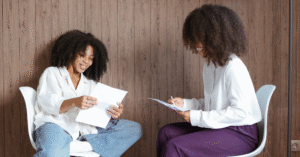 Two black women sit in white chairs, holding papers. The one on the left wears a white shirt and blue jeans and the one on the right wears a white shirt and purple pants.