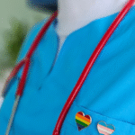 an extreme closeup of someone in a light blue nurse outfit, a pride and trans flag heart pin on the left lapel and a stethoscope