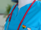 an extreme closeup of someone in a light blue nurse outfit, a pride and trans flag heart pin on the left lapel and a stethoscope