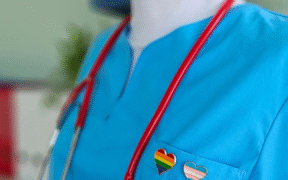 an extreme closeup of someone in a light blue nurse outfit, a pride and trans flag heart pin on the left lapel and a stethoscope