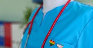 an extreme closeup of someone in a light blue nurse outfit, a pride and trans flag heart pin on the left lapel and a stethoscope