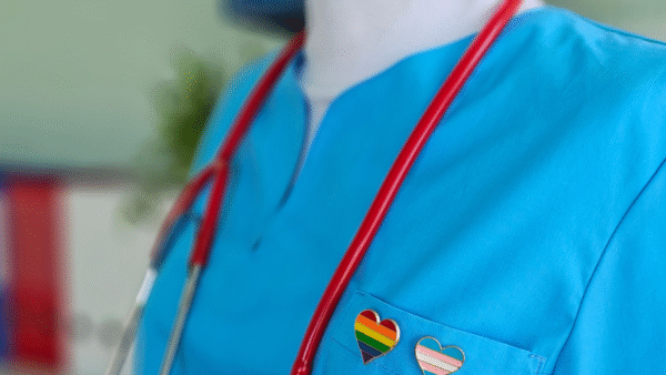 an extreme closeup of someone in a light blue nurse outfit, a pride and trans flag heart pin on the left lapel and a stethoscope