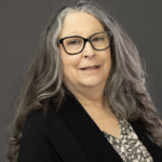 an older white woman with grey-white hair and glasses wearing a black suit jacket over a black and white blouse