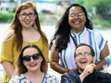 four people stand. Top left is a young white woman with long red hair and glasses wearing a yellow top. Top right is a brown woman with long black hair, glasses, and a pierced nose, wearing a striped short-sleeve shirt. Bottom left is a young white woman with brown hair, wearing sunglasses and a white top with circle patterns, and waving a pride flag. Bottom right is a white man with glasses and a pierced nose wearing a blue flannel top over a black t-shirt and also waving a pride flag