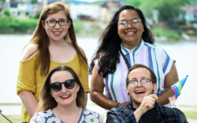 four people stand. Top left is a young white woman with long red hair and glasses wearing a yellow top. Top right is a brown woman with long black hair, glasses, and a pierced nose, wearing a striped short-sleeve shirt. Bottom left is a young white woman with brown hair, wearing sunglasses and a white top with circle patterns, and waving a pride flag. Bottom right is a white man with glasses and a pierced nose wearing a blue flannel top over a black t-shirt and also waving a pride flag