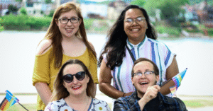 four people stand. Top left is a young white woman with long red hair and glasses wearing a yellow top. Top right is a brown woman with long black hair, glasses, and a pierced nose, wearing a striped short-sleeve shirt. Bottom left is a young white woman with brown hair, wearing sunglasses and a white top with circle patterns, and waving a pride flag. Bottom right is a white man with glasses and a pierced nose wearing a blue flannel top over a black t-shirt and also waving a pride flag