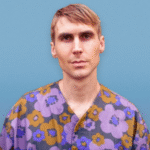 a young white man with short red hair wearing a purple and marigold patterned top