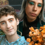 two white people of unknown gender sit on a set of brightly colored steps. The one on the left has brown hair and is wearing a light blue patterned top. The one on the right has long silver-blond hair and is wearing a black turtleneck