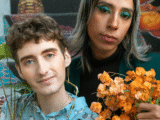 two white people of unknown gender sit on a set of brightly colored steps. The one on the left has brown hair and is wearing a light blue patterned top. The one on the right has long silver-blond hair and is wearing a black turtleneck