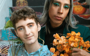 two white people of unknown gender sit on a set of brightly colored steps. The one on the left has brown hair and is wearing a light blue patterned top. The one on the right has long silver-blond hair and is wearing a black turtleneck