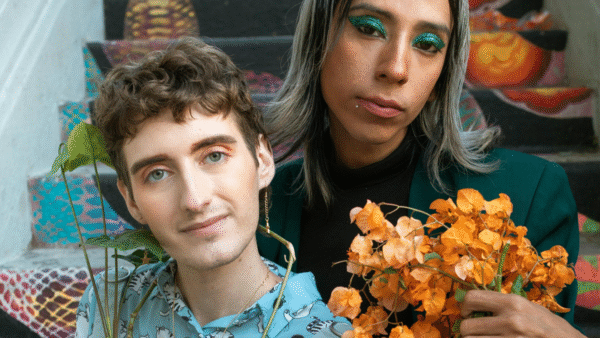 two white people of unknown gender sit on a set of brightly colored steps. The one on the left has brown hair and is wearing a light blue patterned top. The one on the right has long silver-blond hair and is wearing a black turtleneck