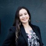 a young woman with long black hair stands, wearing dangling earrings, a black suit jacket, and a top with a black and white leaf pattern
