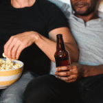 a couple sitting on the couch. The one on the left is brown, wearing a black short-sleeve top and jeans. They are holding a beer bottle and eating popcorn. The one on the right is black, wearing a grey polo and black jeans. They are also holding a beer