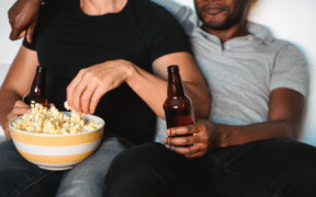 a couple sitting on the couch. The one on the left is brown, wearing a black short-sleeve top and jeans. They are holding a beer bottle and eating popcorn. The one on the right is black, wearing a grey polo and black jeans. They are also holding a beer