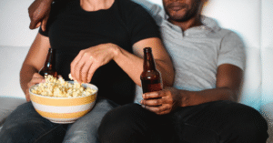 a couple sitting on the couch. The one on the left is brown, wearing a black short-sleeve top and jeans. They are holding a beer bottle and eating popcorn. The one on the right is black, wearing a grey polo and black jeans. They are also holding a beer