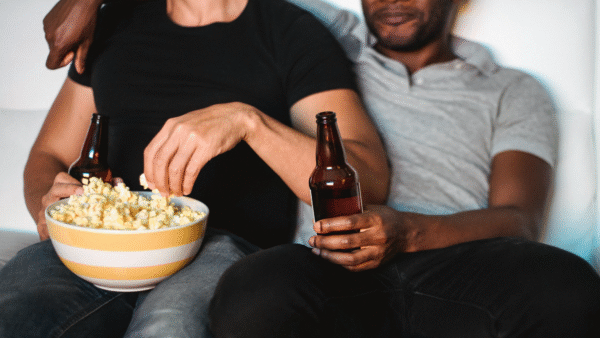a couple sitting on the couch. The one on the left is brown, wearing a black short-sleeve top and jeans. They are holding a beer bottle and eating popcorn. The one on the right is black, wearing a grey polo and black jeans. They are also holding a beer